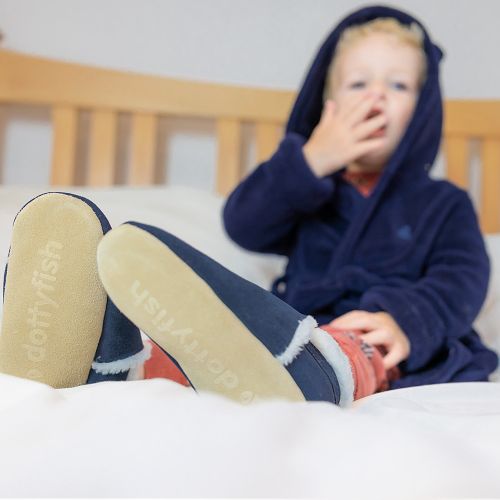Small child sitting in bed, wearing navy suede Dotty Fish barefoot slippers, with fleece lining.