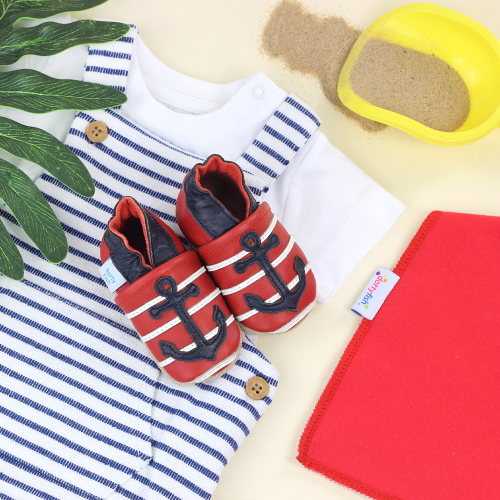 Red leather baby shoes with navy anchor, sitting with baby clothing.