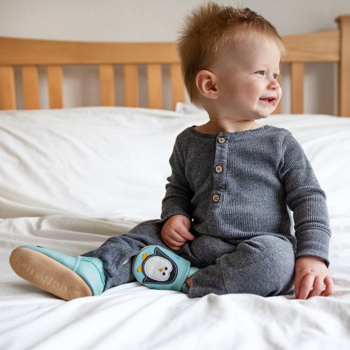 Baby boy sitting on bed, wearing mint green Dotty Fish shoes with white and black penguin design.