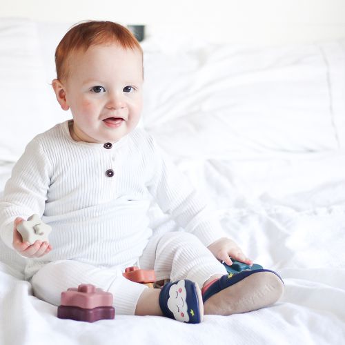 Toddler sitting wearing blue Dotty Fish barefoot shoes with pink ankle trim and white cloud design.