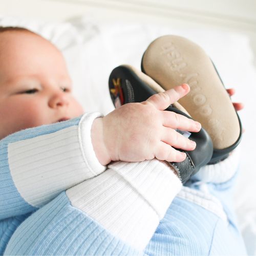 Baby boy lying on bed, wearing charcoal Dotty Fish shoes with silver space rocket design.