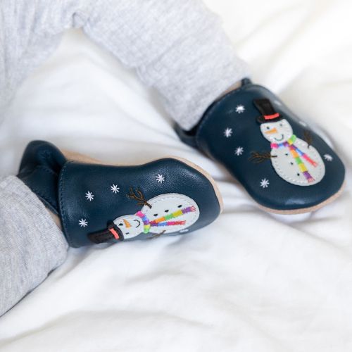 Baby boy sitting, wearing navy Dotty Fish barefoot shoes with white snowman wearing black hat and embroidered rainbow scarf design.