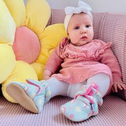 Baby girl wearing pale blue cotton Dotty Fish boots with pink fleece lining and rainbow pattern, sitting with flower cushion.