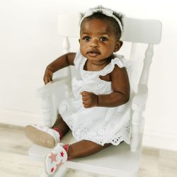 Toddler wearing white leather Dotty Fish first walker barefoot shoes with pink stars, sitting on chair.