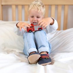 Small boy sitting on bed, wearing navy Dotty Fish shoes with red fire engine design, playing with toy fire engine.