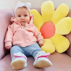 Baby girl wearing rainbow booties, sitting with yellow flower cushion.