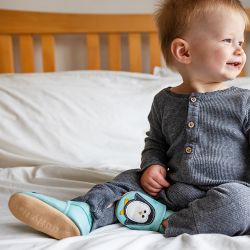 Baby boy sitting on bed, wearing mint green Dotty Fish shoes with white and black penguin design.