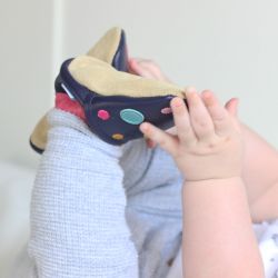 Baby wearing navy Dotty Fish barefoot shoes with pink ankle trim and multicoloured spots.