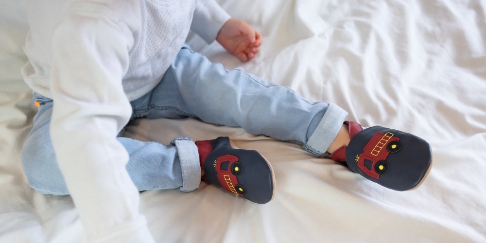 Toddler boy sitting on bed, wearing navy and red fire engine shoes.